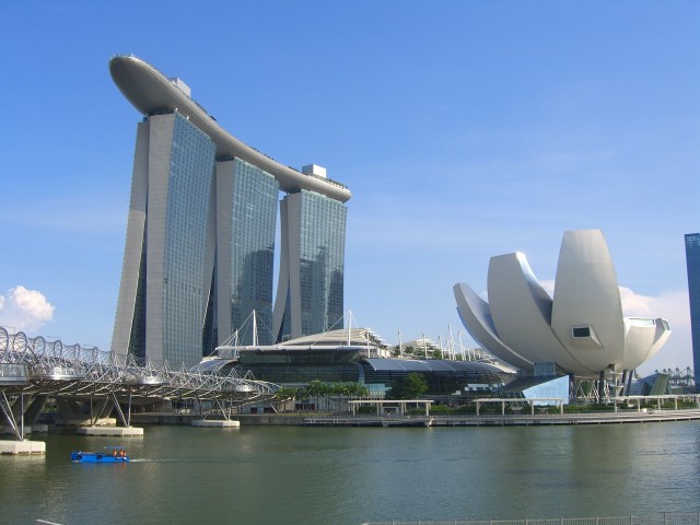 A giant ship sits on top of three sky scrapers, adjacent to a river and a giant building shaped like a half-plucked flower