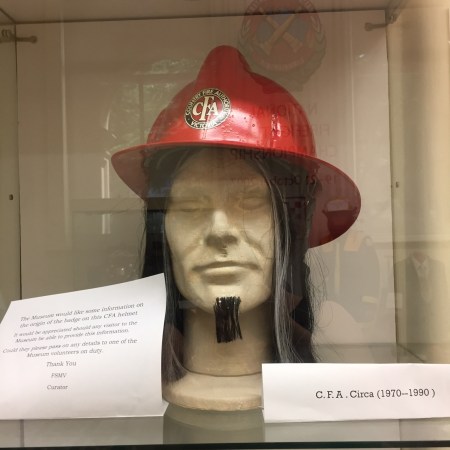 A mannequin's head models a fireman's hat. The face is detailed but unpainted. He wears a long, greying wig, and a small beard that looks like it has been sticky-taped to his face.