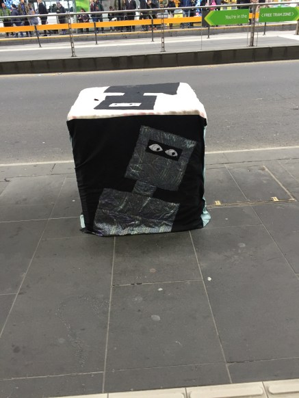 A bollard covered with a fitted quilt depicting Ned Kelly. The top surface has a black Kelly on a white background; the front-facing side has a silver Kelly on a black background with applique eyes. 